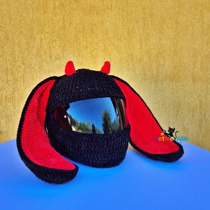 Könnte beinhalten: Ein schwarzer Häkel-Helmüberzug mit zwei kleinen roten Hörnern oben und zwei langen, breiten, herabhängenden Ohren mit roter Innenseite. Der Überzug ist für einen Helm mit dunklem, reflektierendem Visier gedacht. Der Text GetMeYarn ist rechts zu sehen.