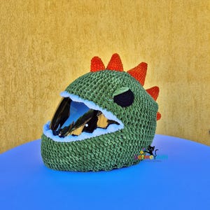 Può includere: Una copertura per casco all'uncinetto verde progettata per sembrare un dinosauro, con una fila di punte arancioni sulla parte superiore, una benda nera sull'occhio e un'apertura bordata di bianco per la visiera. Il testo GetMeYarn è stampato in lettere colorate.