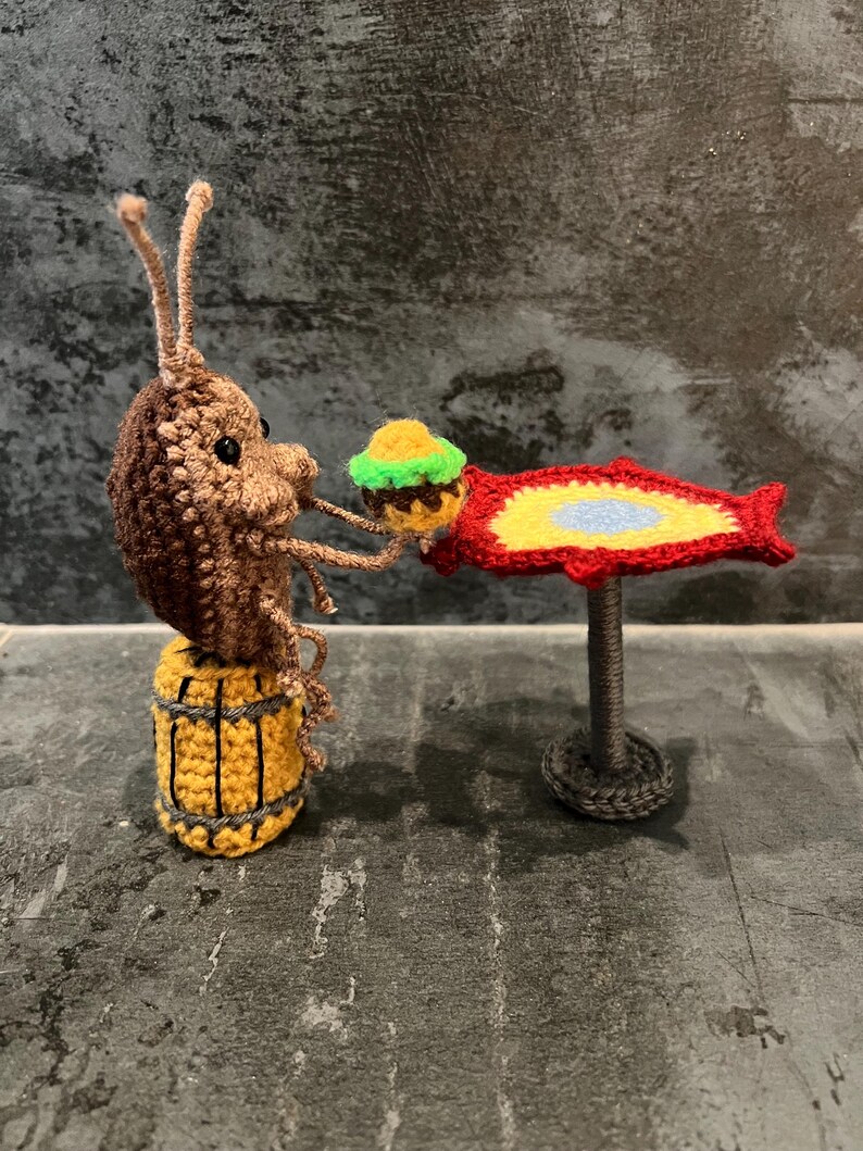 May include: A crocheted brown and tan bug holding a green and yellow crocheted burger. The bug is sitting on a yellow and brown crocheted barrel. The bug is reaching towards a red crocheted fish with a yellow and blue center. The fish is sitting on a gray crocheted stand. Crochet Roach Eating a Burger PDF Crochet Pattern ONLY Digital Download SpongeBob roach pattern