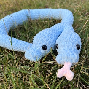 May include: A light blue crocheted snake toy with black eyes and a pink tongue, lying in green grass. The plush snake is made of soft, textured yarn.