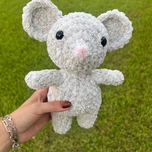 Pode incluir: Um brinquedo de pelúcia cinza feito à mão, em forma de rato, com orelhas grandes e arredondadas, olhos pretos de botão e um nariz rosa. O brinquedo macio e texturizado tem braços e pernas pequenos.