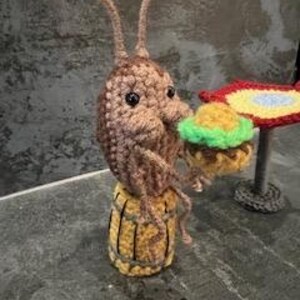 May include: A crocheted brown bug with large eyes and long antennae sitting on a yellow crocheted barrel holding a green and yellow crocheted hamburger. The bug is sitting at a table with a red, yellow, and blue crocheted table cloth.Crochet Roach Eating a Burger PDF Crochet Pattern ONLY Digital Download SpongeBob roach pattern