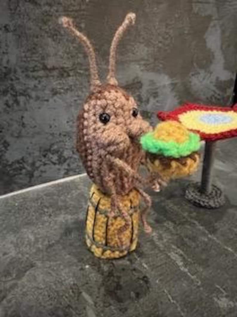 May include: A crocheted brown bug with large eyes and long antennae sitting on a yellow crocheted barrel holding a green and yellow crocheted hamburger. The bug is sitting at a table with a red, yellow, and blue crocheted table cloth.Crochet Roach Eating a Burger PDF Crochet Pattern ONLY Digital Download SpongeBob roach pattern