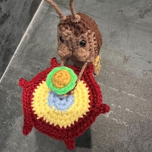 Peut inclure: Un insecte en crochet marron et beige tenant un cercle en crochet rouge, jaune, bleu et vert.