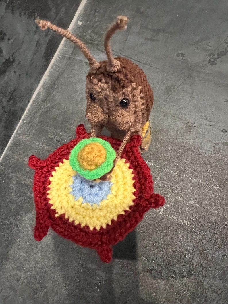 Peut inclure: Un insecte en crochet marron et beige tenant un cercle en crochet rouge, jaune, bleu et vert.