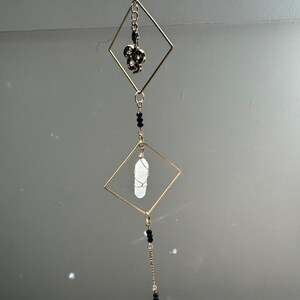 May include: A gold-toned metal hanging mobile with three diamond-shaped frames. The top frame has a black snake charm, the middle frame has a clear quartz crystal, and the bottom frame has a clear crystal teardrop pendant. Black beads are strung on the chain connecting the frames.