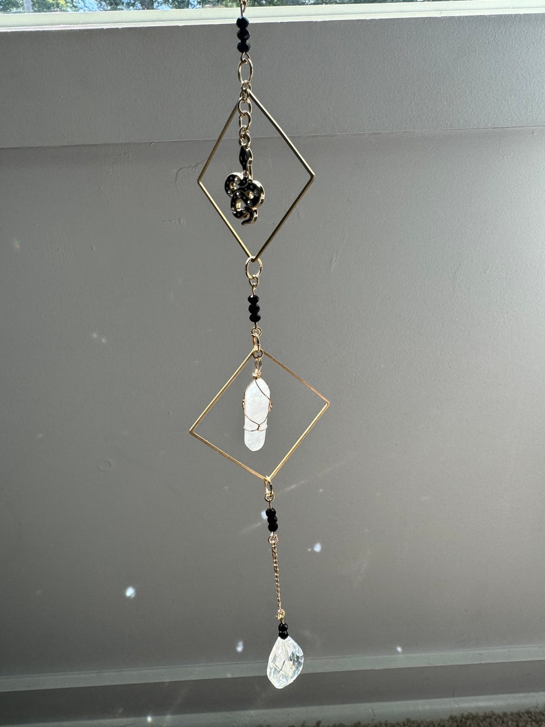 May include: A gold-toned metal hanging mobile with three diamond-shaped frames. The top frame has a black snake charm, the middle frame has a clear quartz crystal, and the bottom frame has a clear crystal teardrop pendant. Black beads are strung on the chain connecting the frames.