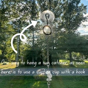 May include: A clear suction cup with a hook attached to a silver chain, hanging from a window. The chain has a crystal prism hanging from it. The background is a blurry view of trees and grass.