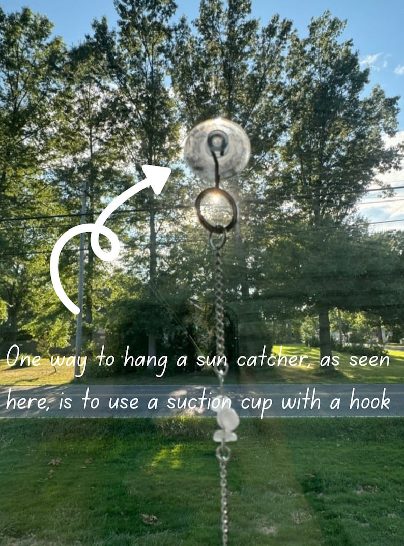 May include: A clear suction cup with a hook attached to a silver chain, hanging from a window. The chain has a crystal prism hanging from it. The background is a blurry view of trees and grass.