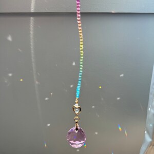 May include: A rainbow crystal suncatcher with a pink crystal ball hanging from a gold chain. The suncatcher is hanging in a window and casting rainbows on the wall.