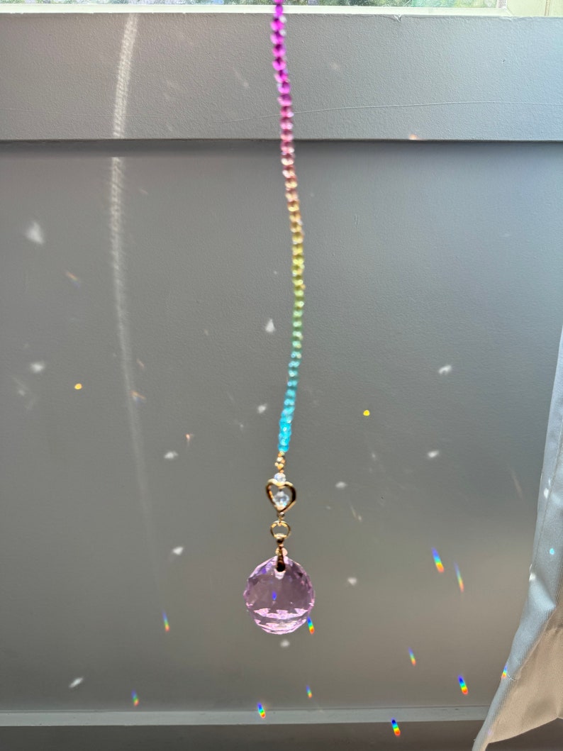 May include: A rainbow crystal suncatcher with a pink crystal ball hanging from a gold chain. The suncatcher is hanging in a window and casting rainbows on the wall.