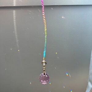 May include: A rainbow crystal suncatcher with a pink crystal hanging from a gold chain. The suncatcher is hanging from a ceiling and casting rainbows on the wall.
