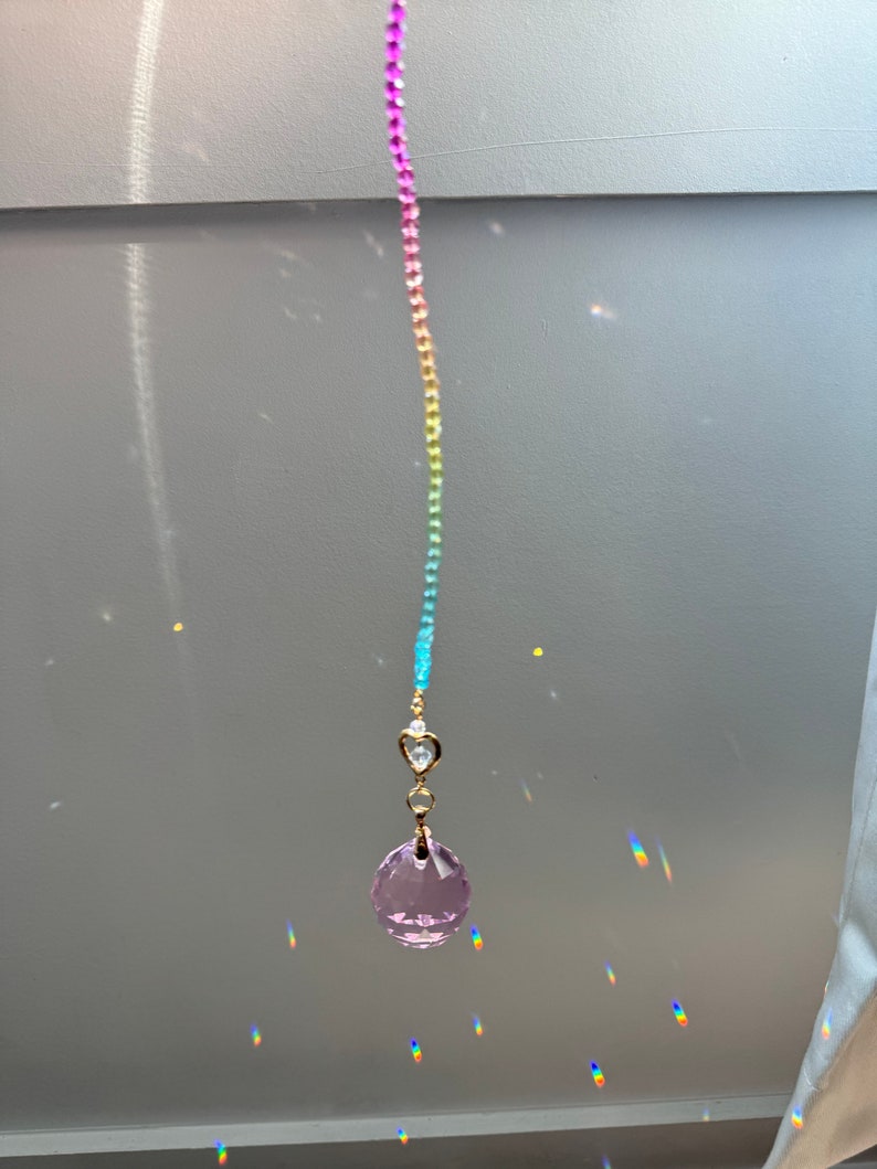 May include: A rainbow crystal suncatcher with a pink crystal hanging from a gold chain. The suncatcher is hanging from a ceiling and casting rainbows on the wall.