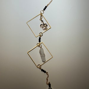 May include: A gold chain with black beads and a gold pendant featuring a snake design. The chain is attached to two gold squares, one of which holds a clear crystal. The chain ends with a teardrop-shaped crystal.