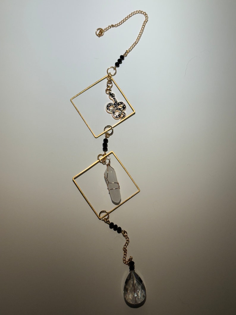May include: A gold chain with black beads and a gold pendant featuring a snake design. The chain is attached to two gold squares, one of which holds a clear crystal. The chain ends with a teardrop-shaped crystal.