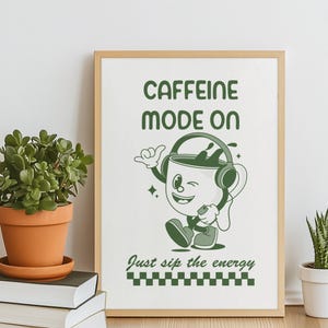 Könnte beinhalten: Gerahmter Druck mit einer Cartoon-Kaffeetasse, die Kopfhörer trägt und zwinkert. Der grüne Text lautet "CAFFEINE MODE ON" und "Just sip the energy". Der Druck befindet sich in einem Holzrahmen.