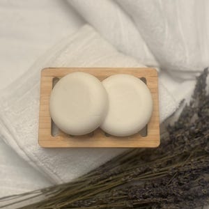 May include: Two round, off-white soap bars rest on a small, rectangular bamboo soap dish. The dish sits on a white towel, with dried lavender sprigs in the foreground. The scene evokes a spa-like atmosphere.