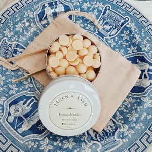 May include: A white tin with a blue and white floral design. The tin contains small, round, white lotion pebbles. The label on the tin reads "LINEN & SASH Lotion Pebbles Sweet Cookie Unscented 2 oz."
