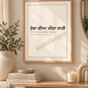 May include: A framed wall art print featuring the Gurmukhi text 'ਤੇਰਾ ਕੀਆ ਮੀਠਾ ਲਾਗੈ', the transliteration 'Tera kiā mīṭhā lāgai', and the English translation 'Your Will is sweet to me'. The print has a thin gold border on a cream background.