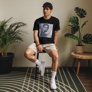 May include: A person wearing a black t-shirt with a graphic of a mouth with a large diamond on the tongue. The person is sitting on a white stool with their legs crossed. They are wearing khaki shorts and white sneakers.