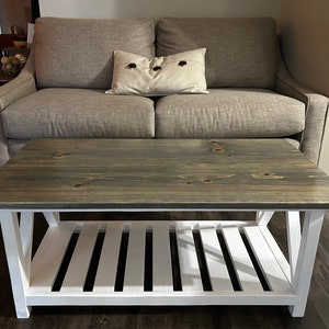 May include: A rectangular coffee table with a gray-stained wooden top and a white slatted shelf. The table is supported by white legs and is positioned in front of a gray sofa. A decorative pillow with dark buttons rests on the sofa.