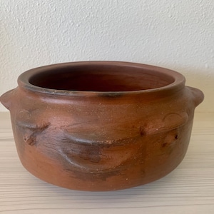 May include: A terracotta pot with two small handles on either side. The pot has a rustic, hand-crafted look and is decorated with a simple design.