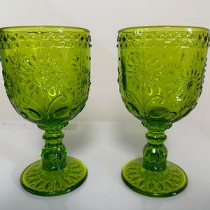 May include: Two matching lime green glass goblets featuring a raised floral pattern on the bowl, a beaded rim detail, and a textured pedestal base. The glassware has a translucent finish with intricate embossed sunflower-like motifs.