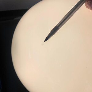 K&ouml;nnte beinhalten: Nahaufnahme einer wei&szlig;en, runden, glatten Oberfl&auml;che mit einem kleinen, dunklen Fleck in der Mitte. Ein Stift zeigt auf den Fleck.