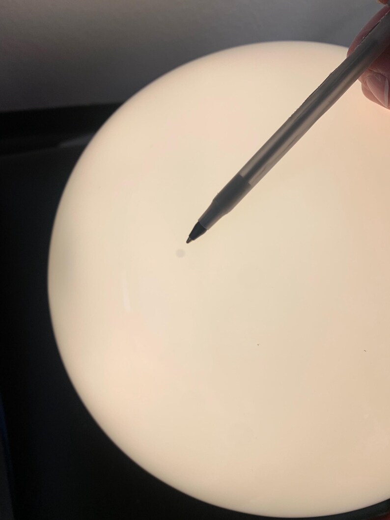 K&ouml;nnte beinhalten: Nahaufnahme einer wei&szlig;en, runden, glatten Oberfl&auml;che mit einem kleinen, dunklen Fleck in der Mitte. Ein Stift zeigt auf den Fleck.