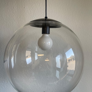 May include: A clear glass globe pendant light with a black metal fixture and a white bulb. The spherical shade hangs from a black cord, creating a modern and minimalist aesthetic. The light fixture is designed for indoor use.