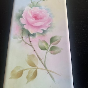 May include: A rectangular painting featuring a pink rose with green leaves and thorns. The rose is the focal point, with delicate brushstrokes and shading. The background is a soft blend of pink and light green, creating a serene and elegant feel.