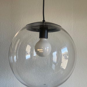 May include: A clear glass globe pendant light with a black metal cap and a white bulb. The light hangs from a black cord, creating a modern and minimalist aesthetic. The globe reflects the surrounding environment.