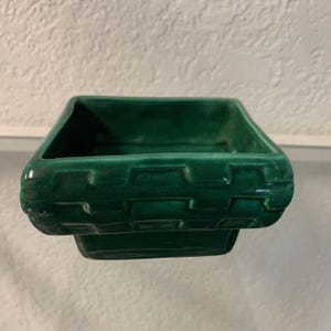 May include: A square, dark green ceramic dish with a basket weave design around the sides. The dish has a glossy finish and a slightly raised base. The interior is smooth and the corners are rounded.