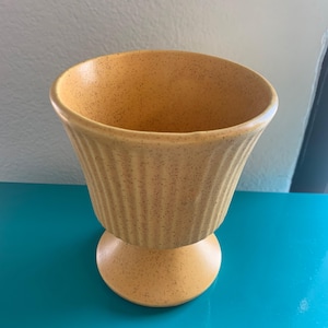 May include: A yellow ceramic vase with a speckled finish. The vase has a flared, ribbed body and sits on a rounded base. The color is a warm, muted yellow, and the overall design is vintage.