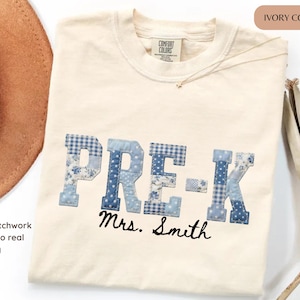 May include: An ivory-colored t-shirt featuring the text PRE-K in a blue and white faux patchwork print with various patterns including floral, gingham, and polka dots. Below the text, the name Mrs. Smith is printed in a black cursive font.