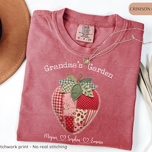 May include: Crimson-colored t-shirt with "Grandma's Garden" embroidered above a faux patchwork strawberry. The shirt includes the names Megan, Sophia, and Emma. A silver necklace and a brown hat are also visible.