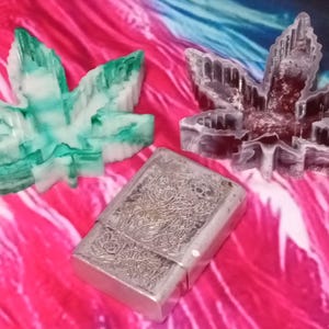 May include: Three decorative cannabis leaf-shaped items in green, white, and maroon hues, alongside a silver rectangular box with an ornate design. The items are arranged on a pink and blue patterned surface.