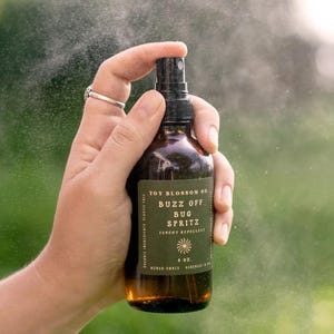 May include: A brown glass bottle of "Buzz Off Bug Spritz" insect repellent is held in a hand. The label is green with white text and a sunburst design. The bottle is spraying a fine mist. The bottle contains 4 oz.