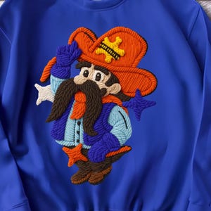 Op de afbeelding: Koning blauwe sweatshirt met een textuur cowboy ontwerp. De cowboy draagt een oranje hoed met een gele ster, een blauwe jas en een bruine snor. Het ontwerp is gemaakt van een garenachtig materiaal.