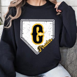 May include: Black sweatshirt featuring a distressed home plate design with a yellow and black letter "G" and the word "Pirates" in a script font. The design has a vintage, worn look.