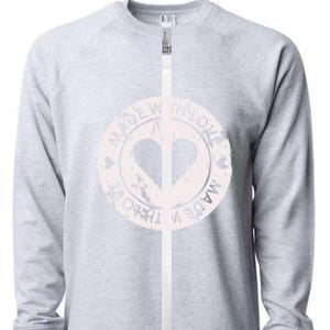 May include: A light gray sweatshirt with long sleeves. The front features a distressed, light pink circular graphic with a heart and the words "MADE WITH LOVE" around it. The sweatshirt has a crew neck.