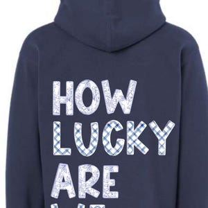 以下が含まれることがあります： 「HOW LUCKY ARE WE」の文字が白で、青い花柄とチェック柄の模様が入ったネイビーブルーのパーカー。フード付きで、柔らかい素材でできています。