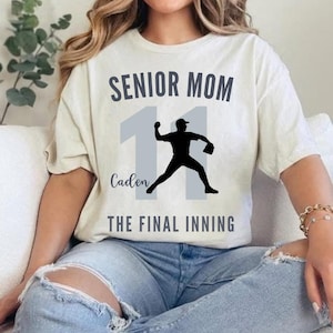 May include: Cream-colored t-shirt with the text "SENIOR MOM" above a baseball player silhouette and the number 11. Below the silhouette is the name "Caden" and the phrase "THE FINAL INNING".