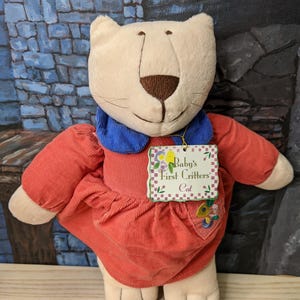 May include: A plush cat toy with a cream-colored head, brown nose, and whiskers. It wears a red corduroy dress with a blue collar. A tag reads "Baby's First Critters Cat."