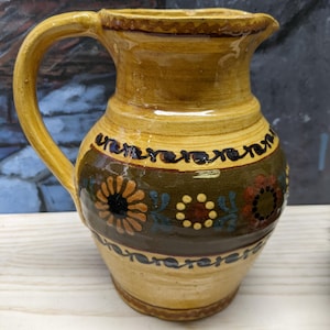 May include: A handcrafted ceramic pitcher with a yellow glaze and a curved handle. The pitcher features a dark green band with floral and dot designs in orange, red, and blue. The rim and handle are also yellow.