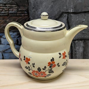 May include: Cream-colored ceramic teapot with a silver rim and lid. The teapot features a floral design with red flowers and black leaves. The handle and spout are curved.