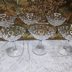 May include: Six clear, stemmed champagne coupe glasses. The glasses have a diamond-cut pattern on the bowl and a round base. The background is a floral tapestry.