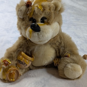 Puede incluir: Un oso de peluche color canela con nariz negra y ojos oscuros grandes. El oso está cubierto con una sustancia amarilla brillante similar a la miel, con dos pequeños tarros de cerámica con la palabra "HONEY" en sus patas y un cazo de madera.