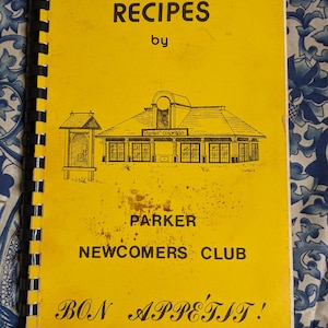 May include: A yellow cookbook with the title "Recipes by Parker Newcomers Club" and the text "Bon Appetit!" printed on the front cover. The cover also features a black and white illustration of a building with the words "Parker Colorado" above the entrance.
