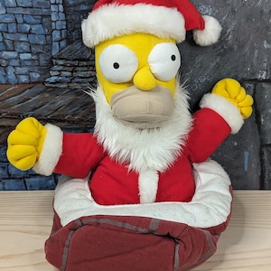 May include: A plush Homer Simpson figure dressed as Santa Claus. The figure has a yellow face, large white eyes, and a beige mouth. He wears a red Santa suit with white trim and a matching hat. The figure is seated in a red sack.